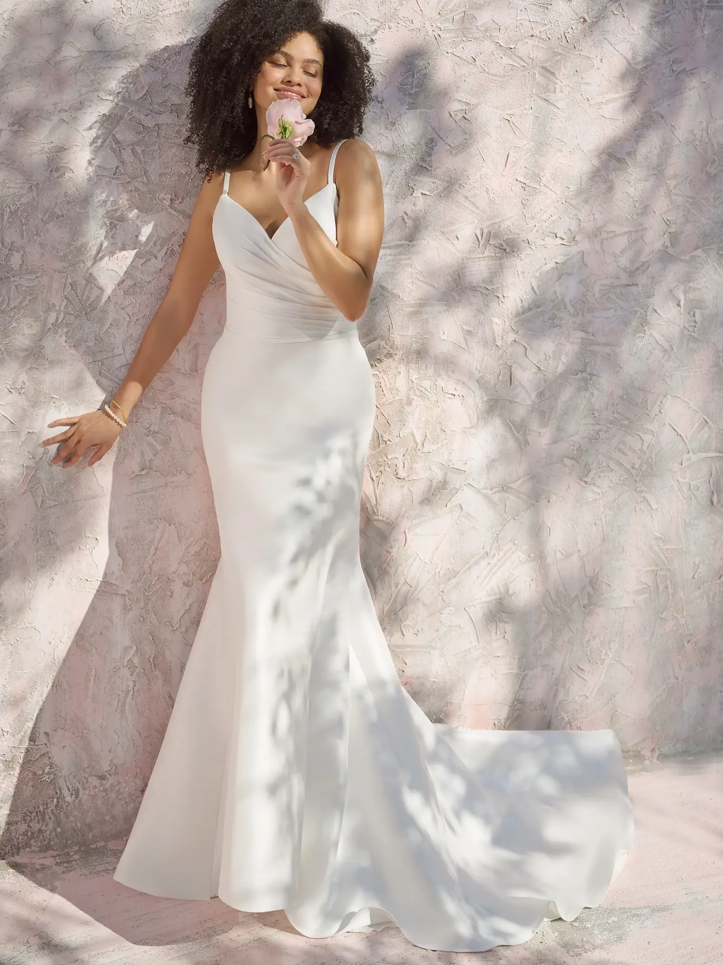 A woman with curly hair wearing a fitted white dress, standing against a light pink textured wall, holding a small flower.