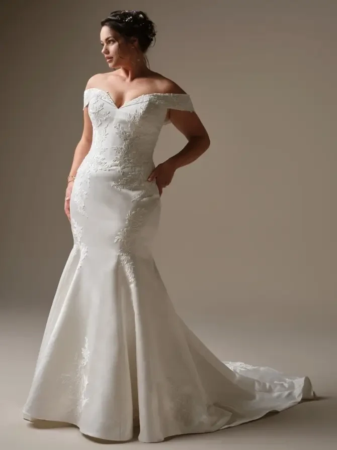 A woman modeling an elegant, off-the-shoulder wedding gown with a fitted bodice and a flowing skirt.
