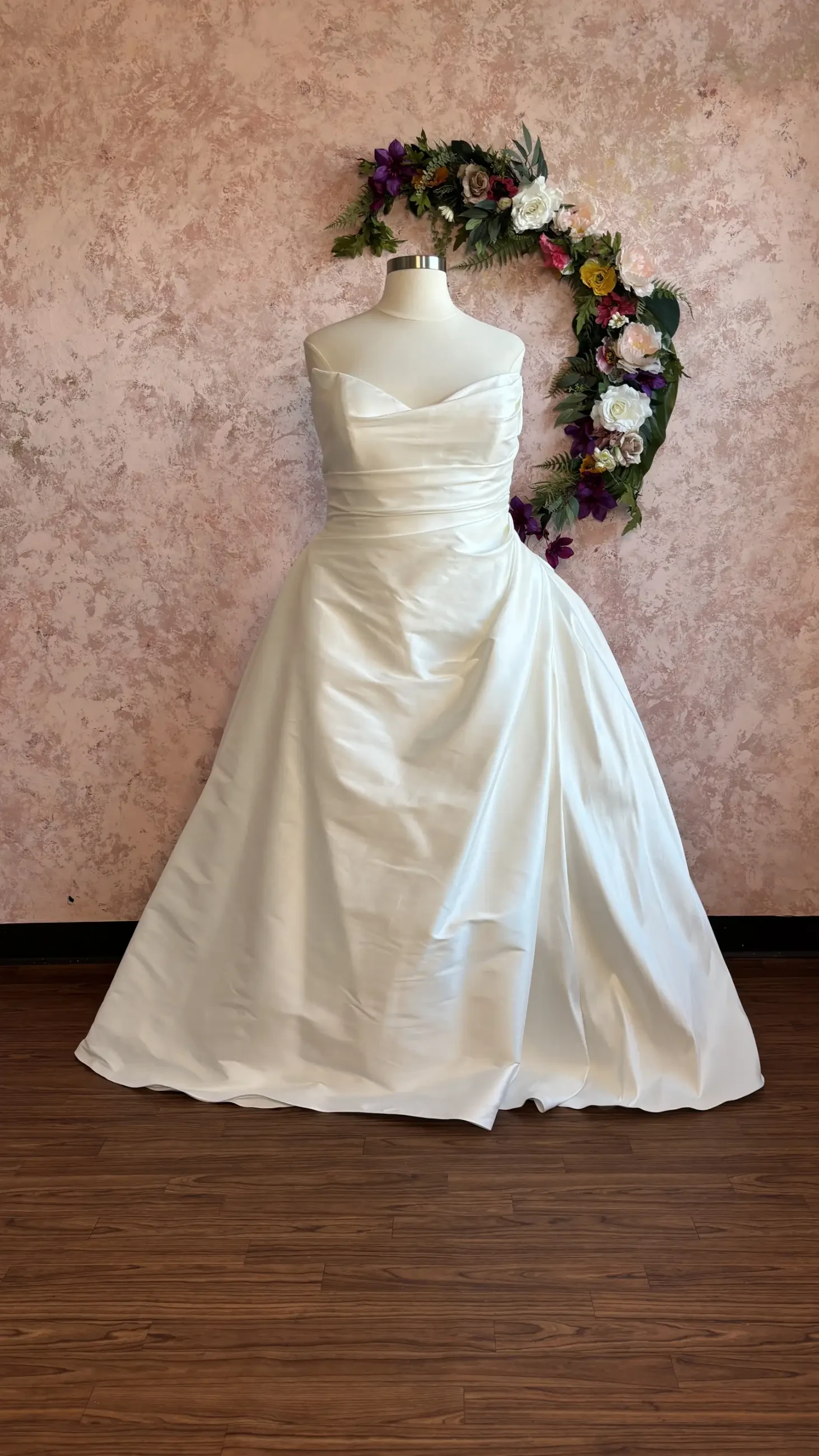 A white wedding dress displayed on a mannequin, with a floral wreath in the background.