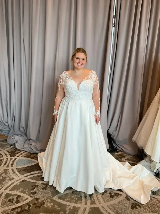 A woman wearing a long-sleeve, white wedding dress stands in front of gray curtains. She is smiling and the dress has intricate lace details on the sleeves.