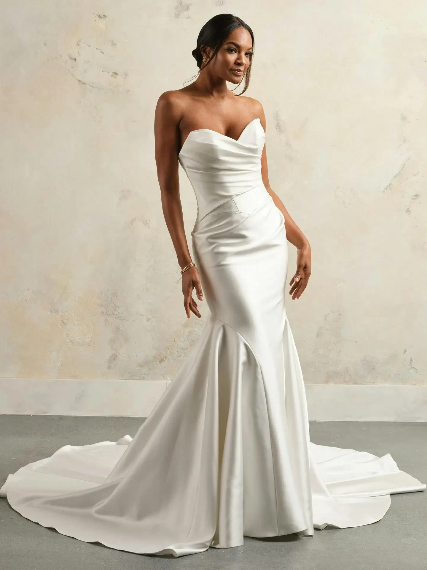 A woman wearing a strapless white wedding gown, posing elegantly with her hands on her hips. The dress has a fitted silhouette and a train, set against a neutral background.