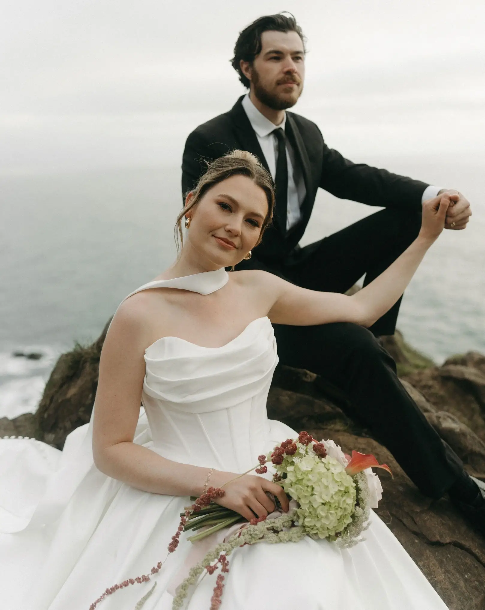 A bride in a white wedding dress holding a bouquet sits on a rock near the ocean, while a groom in a black suit sits behind her.
