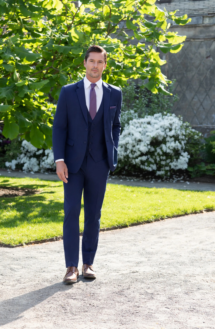 A man in a navy suit with a tie, standing in a garden with green foliage and white flowers.