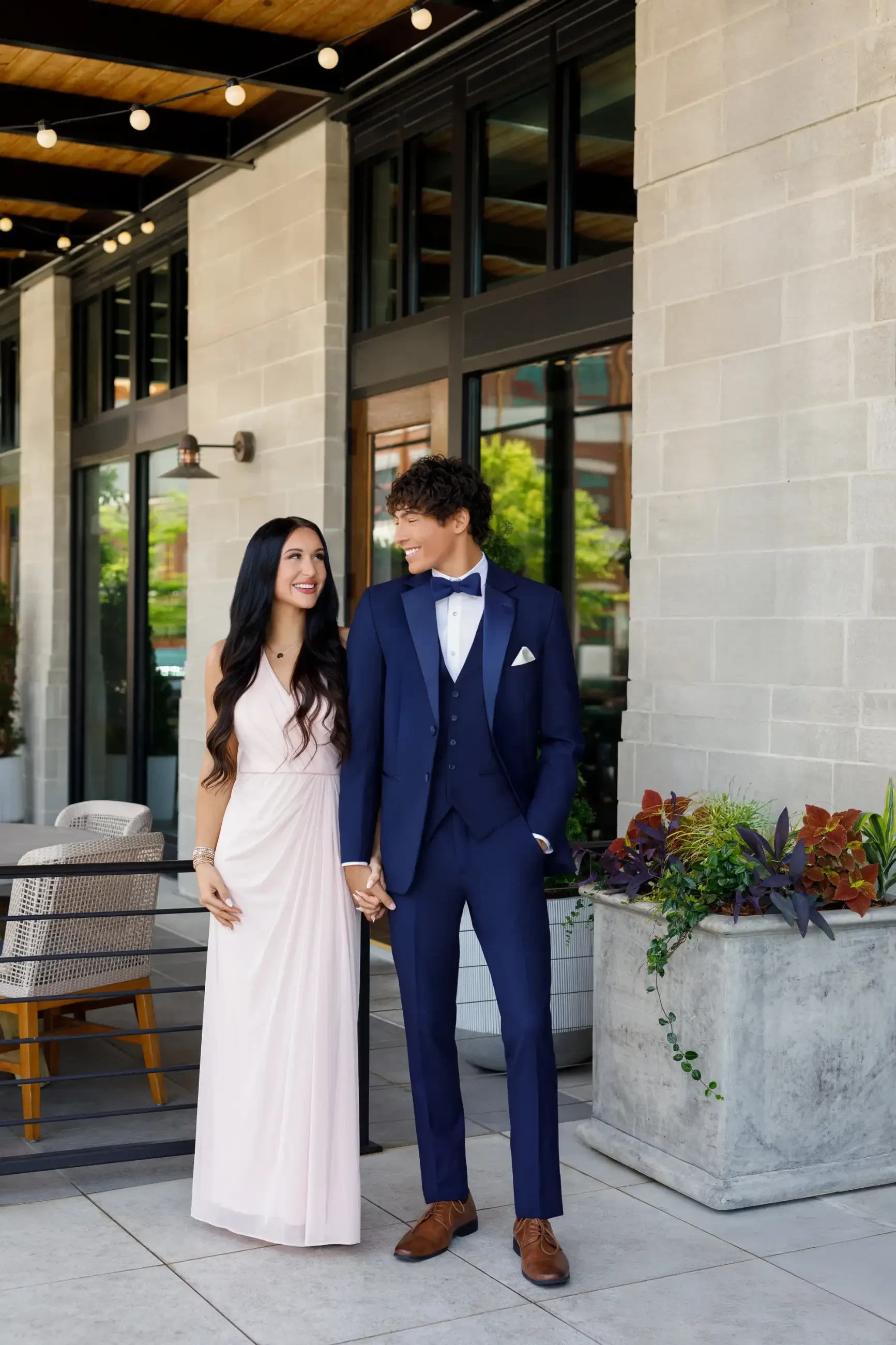 A couple standing outside a building, the woman in a light pink dress and the man in a dark suit with a bow tie, both holding hands and smiling at each other.