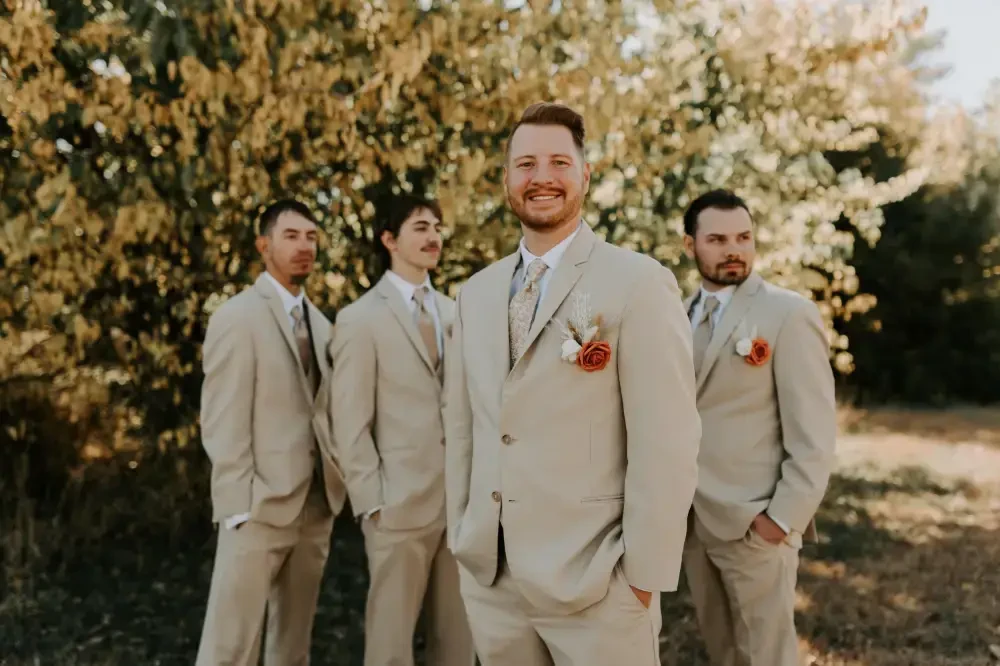 A group of four men in light beige suits standing outdoors, with the man in front smiling and others looking away. Each has a rose boutonni&egrave;re on their lapel, with trees in the background.