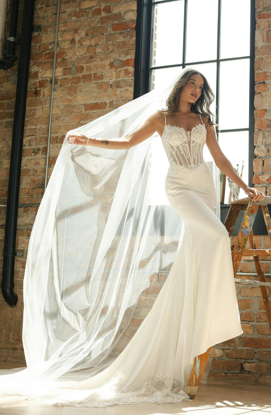 A woman in a bridal gown posing with a flowing veil in an industrial-style room with brick walls and large windows.