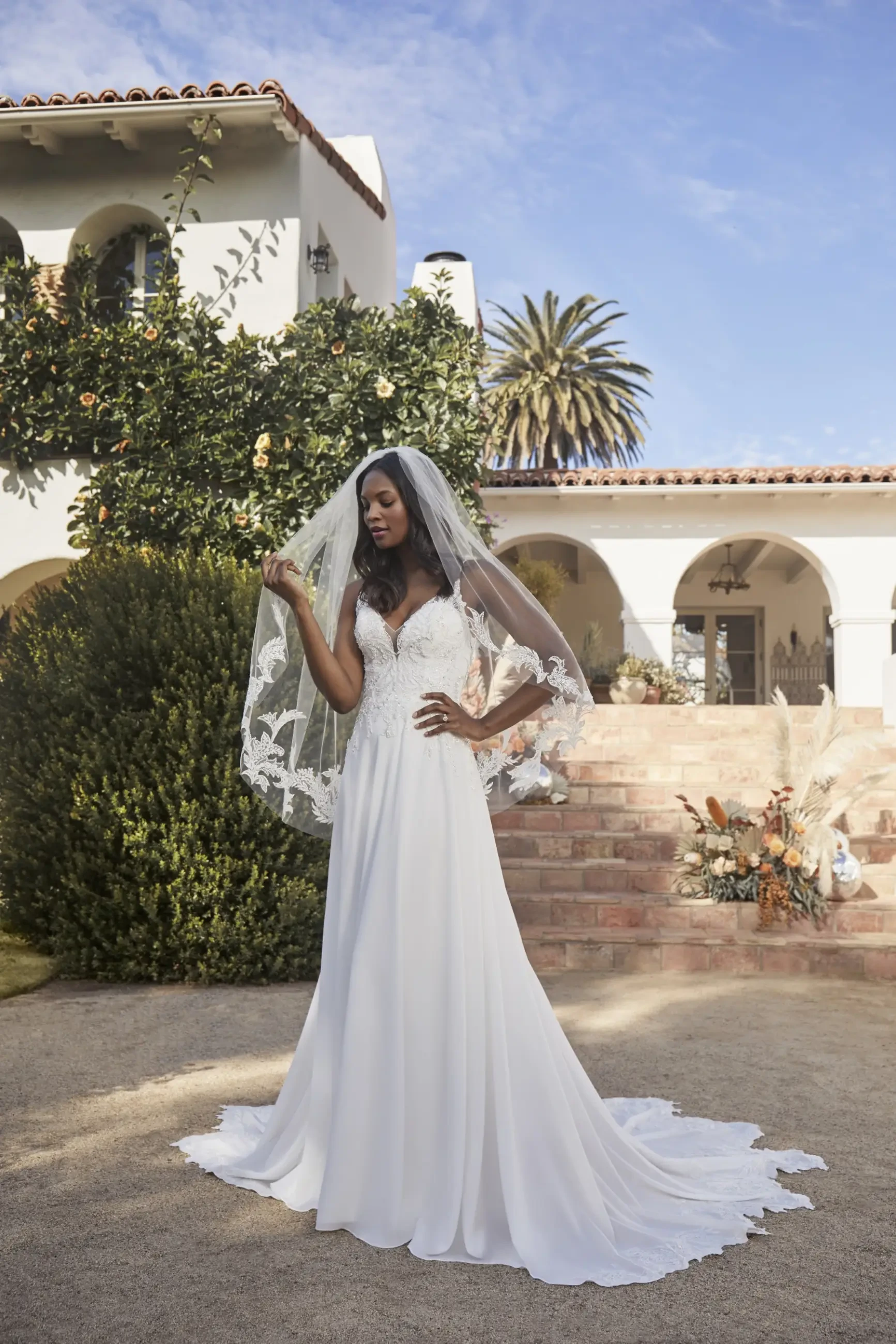 A woman in a white wedding dress and veil stands outdoors, with palm trees and a building in the background.